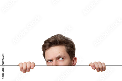 Young man with surprised eyes peeking out from behind billboard paper poster. Man peeking out from the edge and looking at camera isolated on a white background