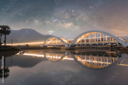 Light from the train that runs through the Tha Chom Phu white bridge over the river at night in the sky with stars and the Milky Way illuminating in the dark. 