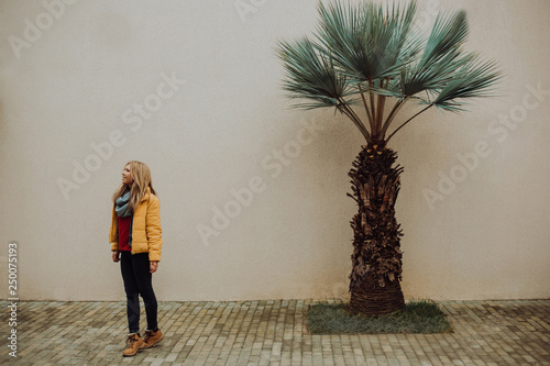 A beautiful young tanned girl stands near a yellow wall in the south. Fashionable dress, bright make-up, tan. Palms, photo