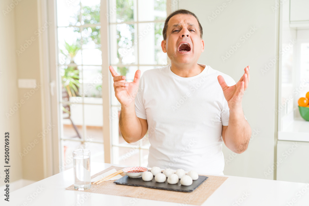 Middle age man eating asian dumplings at home crazy and mad shouting and yelling with aggressive expression and arms raised. Frustration concept. Middle age man eating asian dumplings at home crazy and mad shouting and yelling with aggressive expression and arms raised. Frustration concept.