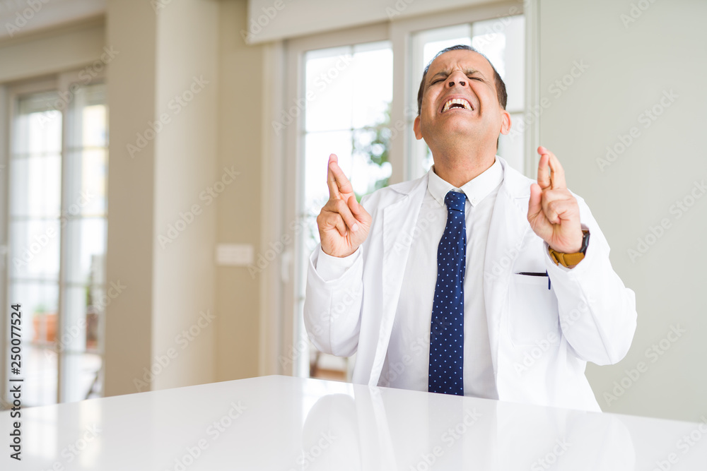 Middle age doctor man wearing medical coat at the clinic smiling crossing fingers with hope and eyes closed. Luck and superstitious concept. Middle age doctor man wearing medical coat at the clinic smiling crossing fingers with hope and eyes closed. Luck and superstitious concept.