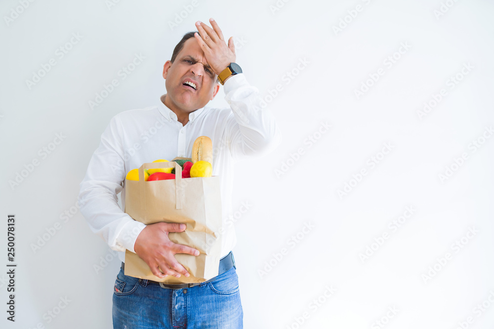 Middle age man holding groceries shopping bag over white background stressed with hand on head, shocked with shame and surprise face, angry and frustrated. Fear and upset for mistake. Middle age man holding groceries shopping bag over white background stressed with hand on head, shocked with shame and surprise face, angry and frustrated. Fear and upset for mistake.