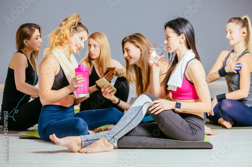 Group of six female sitting on the floor in yoga, pilates studio, having break talking, relaxing, drink water. Healthy lifestyle, teamwork, training, workout, friendship concept.