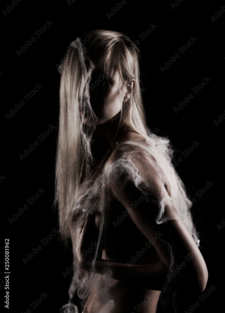 Foto de Beautiful topless blonde girl with hair falling on her face, stands  sideways covered with spider web, with a her hand covers her big boobs.  Black background. Artistic noir silhouette photo