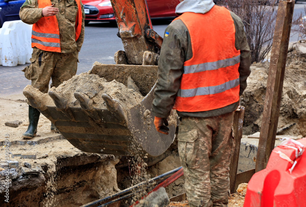 A bucket of a heavy construction excavator surrounded by road workers digs a trench when repairing a city sewer on the roadway. A bucket of a heavy construction excavator surrounded by road workers digs a trench when repairing a city sewer on the roadway.