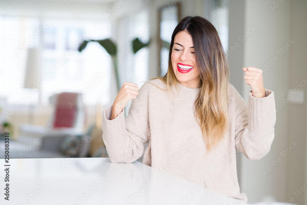 Young beautiful woman wearing winter sweater at home very happy and excited doing winner gesture with arms raised, smiling and screaming for success. Celebration concept.