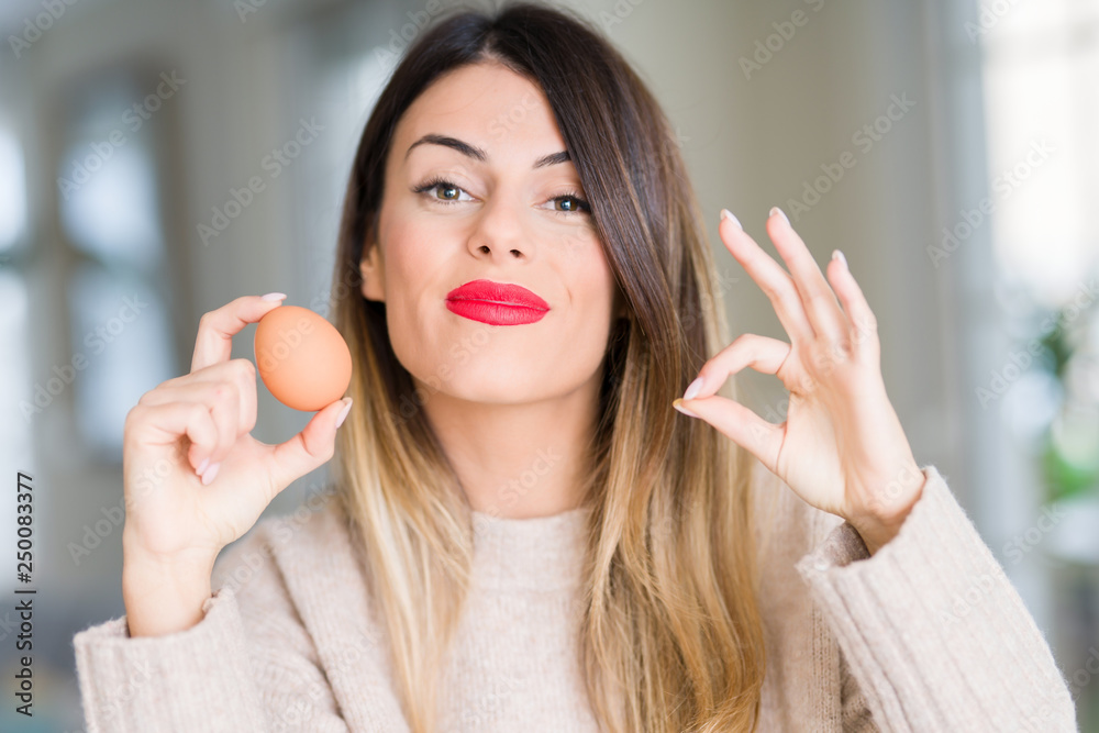 Young beautiful woman holding fresh egg at home doing ok sign with fingers, excellent symbol Young beautiful woman holding fresh egg at home doing ok sign with fingers, excellent symbol