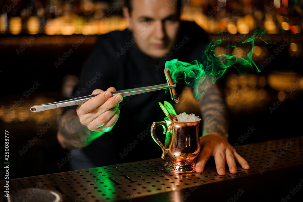 Bartender adding to the cocktail with dried orange a smoked cinnamon with tweezers in the red light