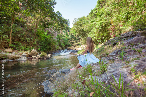 Young woman with long hair sitting back on the rocks near small river with clean water among green tropical trees and plants while sky is blue on the north of Bali island  Indonesia