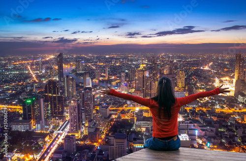 Photography Rear view of female traveler with raised hands standing on the top of a tower and looking at the  urban city with sunset sky