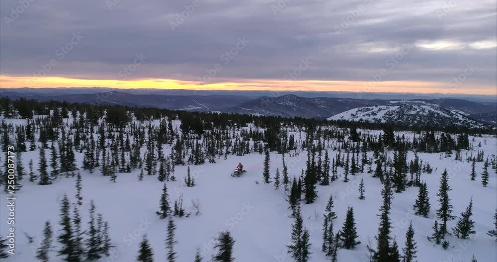 a guy on a mountain snowmobile rides on the background of a beautiful sunrise in a forest mountain valley with large fields of snow. movement on a snowmobile in the mountains, the camera flies behind a guy on a mountain snowmobile rides on the background of a beautiful sunrise in a forest mountain valley with large fields of snow. movement on a snowmobile in the mountains, the camera flies behind