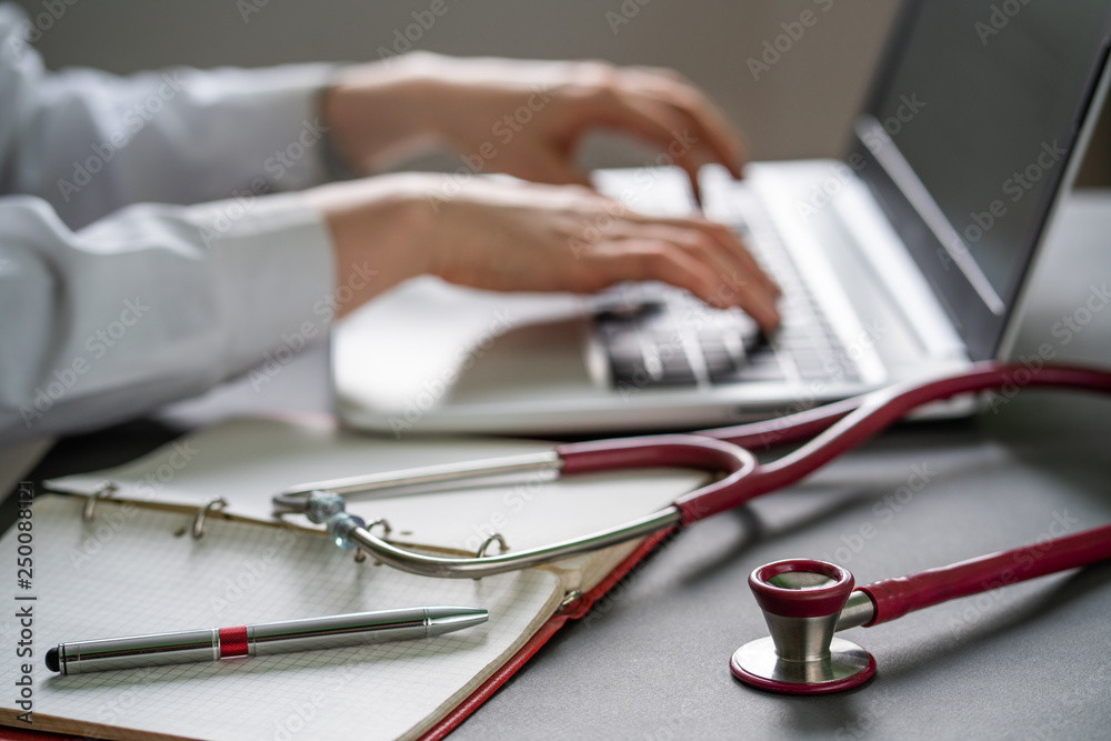 Stethoscope and background doctor using laptop at desk in clinic
