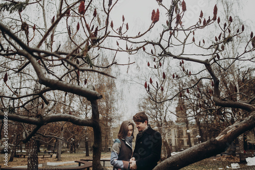 romantic stylish couple hugging gently, holding hands in autumn park under unusual tree. man and woman embracing. family togetherness concept. calm atmospheric moment