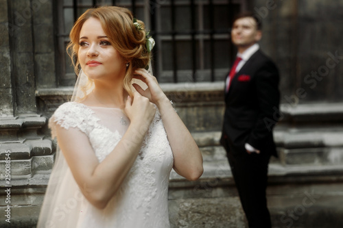 Beautiful blonde bride in luxury white wedding dress posing outdoors in italian street, face closeup of gorgeous newlywed woman, groom standing in the background © sonyachny