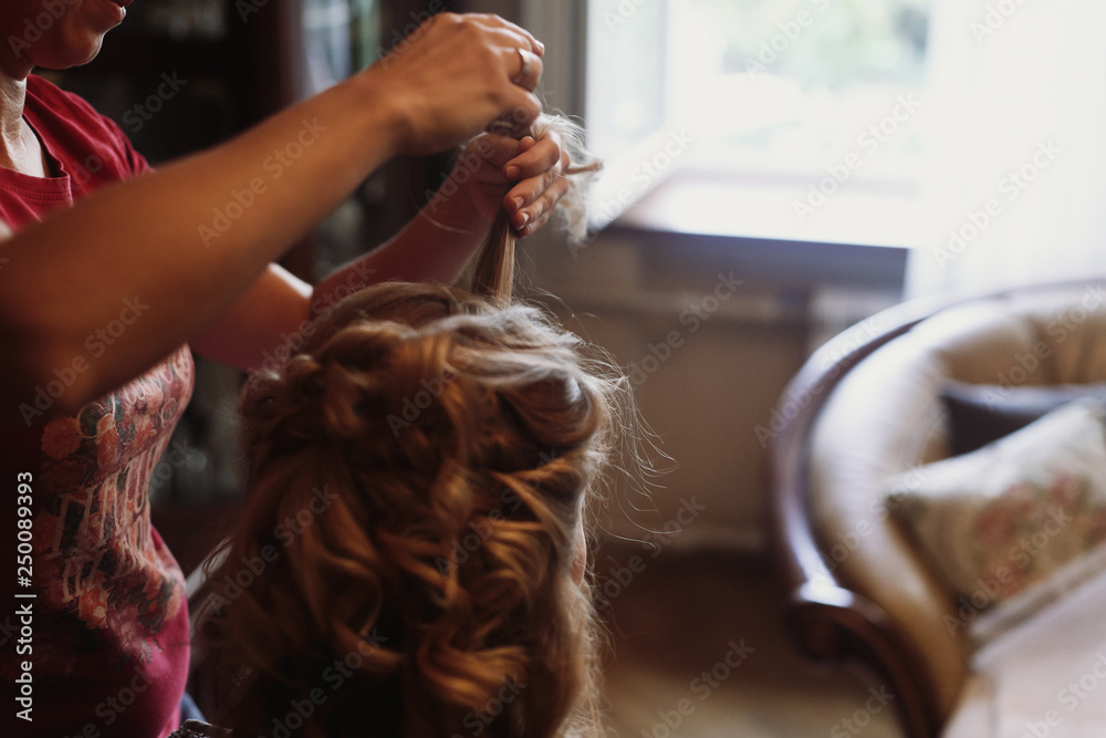 Naklejka premium Professional stylist doing hairstyle for beautiful woman, blonde bride getting her hair done by hairdresser, morning wedding preparation closeup