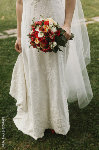 Beautiful bride holding wedding bouquet, stylish bride in lace white dress holding red roses bouquet in her hand closeup, posing outdoors near church