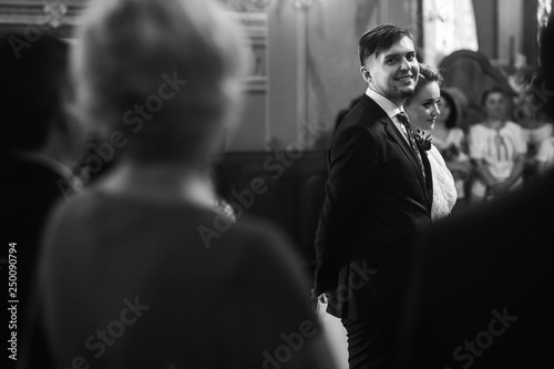Handsome groom looking at beautiful bride, happy groom in stylish black suit glancing at gorgeous blonde bride in church during wedding ceremony face closeup