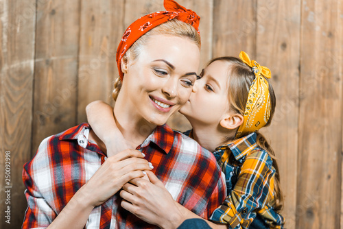 cute daughter kissing cheek of happy mother near wooden fence