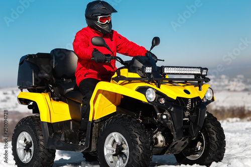 Photo of attractive young man in red warm winter clothes and black helmet on the ATV 4wd quad bike stand in heavy snow with deep wheel track. Moto winter sports.