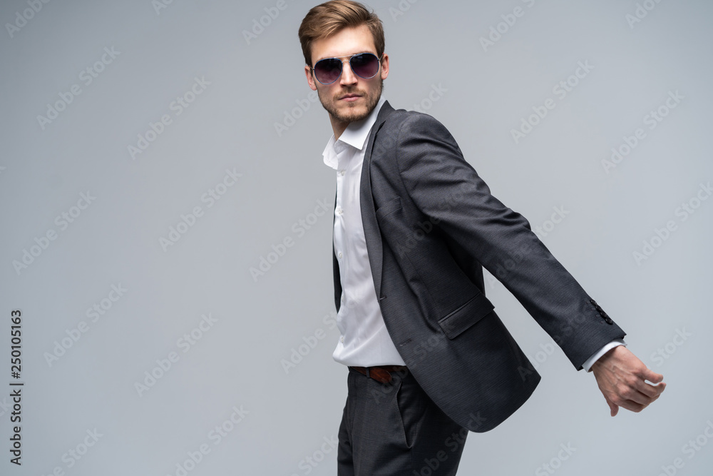 Feeling free and comfortable in his style. Handsome young man in full suit and sunglasses moving in front of gray background. Feeling free and comfortable in his style. Handsome young man in full suit and sunglasses moving in front of gray background.