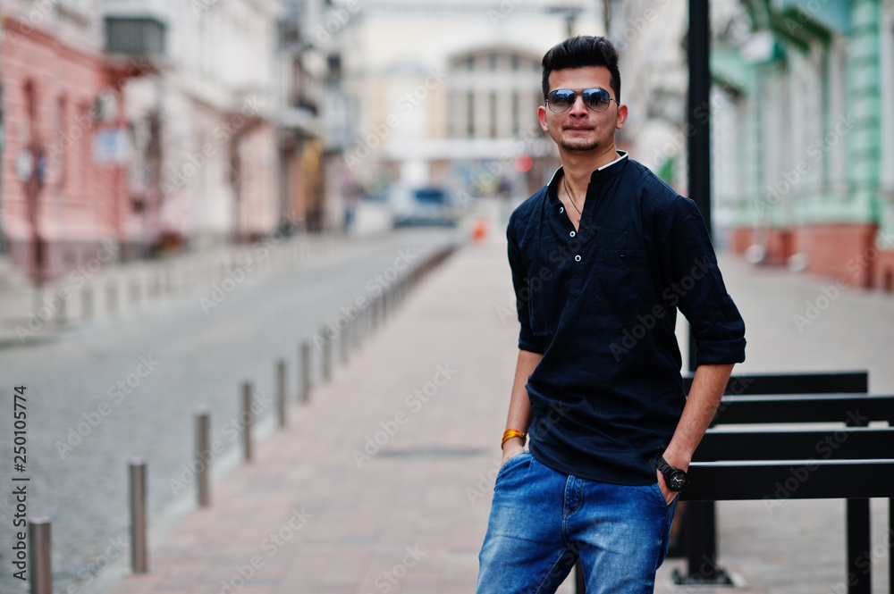Stylish indian model man in casual clothes, black shirt and sunglasses posed outdoor at street of India.