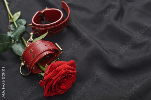 Erotic toys. Top view of red leather handcuffs and rose against of black silk fabric.