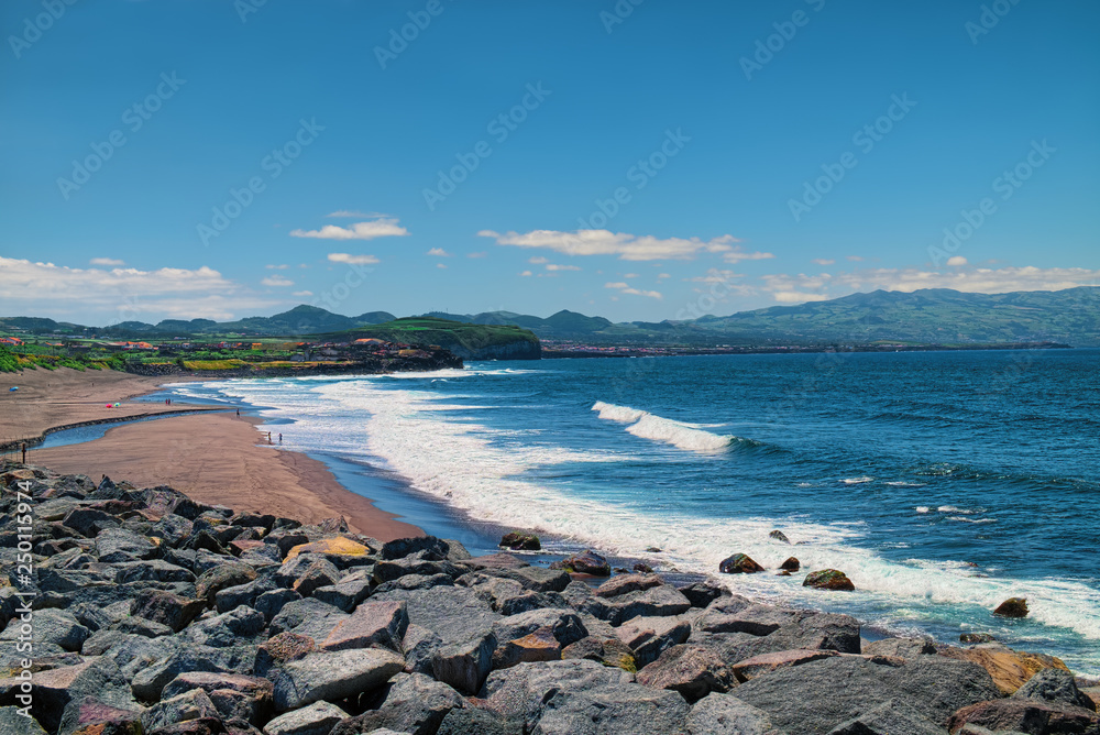 Oceanic coast of Sao Miguel island of Azores, Portugal, near the Ribeira Grande town, with mountains visible on skyline and sand beach on left side.