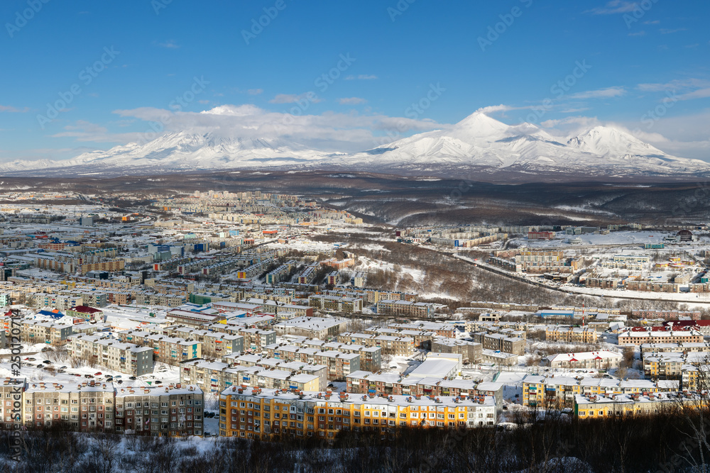 Winter landscape residential buildings of Petropavlovsk-Kamchatsky City, scenery volcanoes of Kamchatka Peninsula: Koryak Volcano, Avacha Volcano, Kozelsky Volcano on clear sunny day with blue sky.