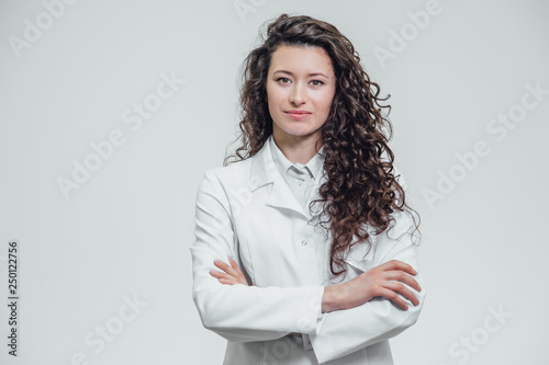 Portrait of happy young smiling girl doctor. Dressed in a white robe. Evenly standing with crossed hands on a gray background.
