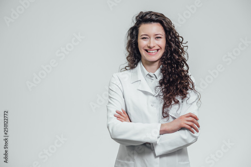 Portrait of happy young smiling girl doctor. Dressed in a white robe. Evenly standing with crossed hands on a gray background.