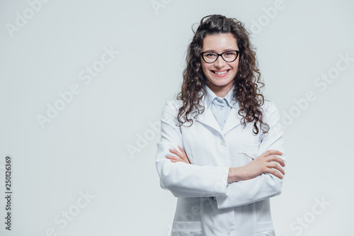 Portrait of smiling young female doctor. Beautiful brunette in white medical gown in glasses. Holding without a stethoscope on a gray background.