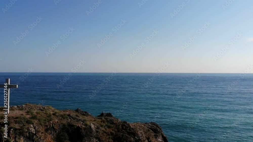 inspiring upper motion over white cross on rock lit by sun to endless blue sea under clear sky in summer inspiring upper motion over white cross on rock lit by sun to endless blue sea under clear sky in summer