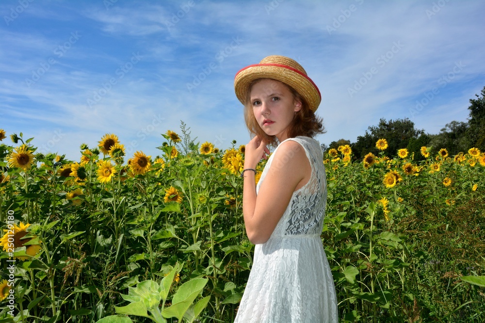 Junge Frau steht in einem Sonnenblumen Feld mit Kleid und Strohhut und hält mit einer Hand ihre Haare