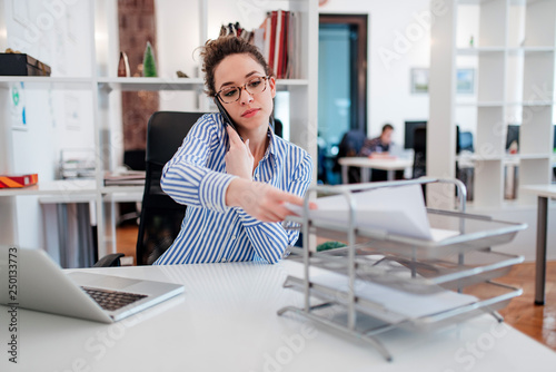 Foto Young lady at desk doing paperwork and talking on smartphone.