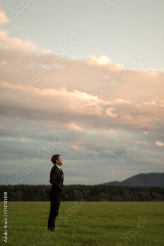 Businessman in a suit standing in green meadow under a glowing evening sky © Gajus