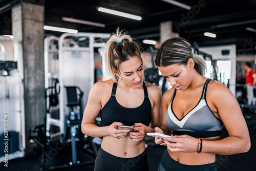 Two female athlete friends using smart phones at the gym.