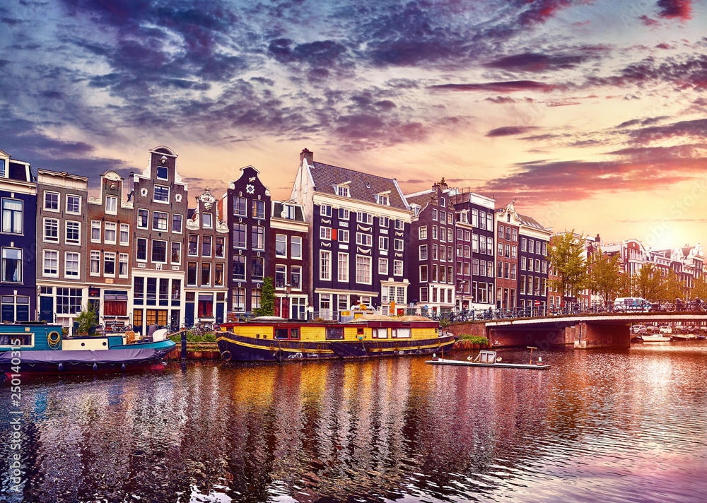 Amsterdam, Netherlands. Floating Houses and houseboats and boats at channels by banks. Traditional dutch dancing houses among trees. Evening autumn street above water pink sunset sky with clouds.