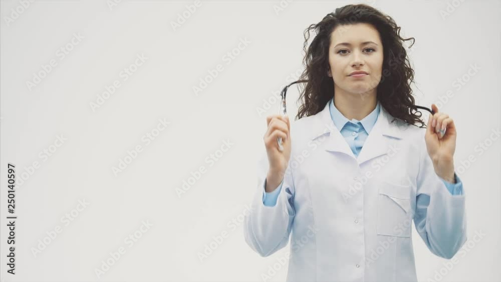 Beautiful young brunette doctor wearing medical clothes on gray background. During this she wears the phonendoscope on her neck. Has beautiful black long hair. Dressed in glasses Slow video. Beautiful young brunette doctor wearing medical clothes on gray background. During this she wears the phonendoscope on her neck. Has beautiful black long hair. Dressed in glasses Slow video.