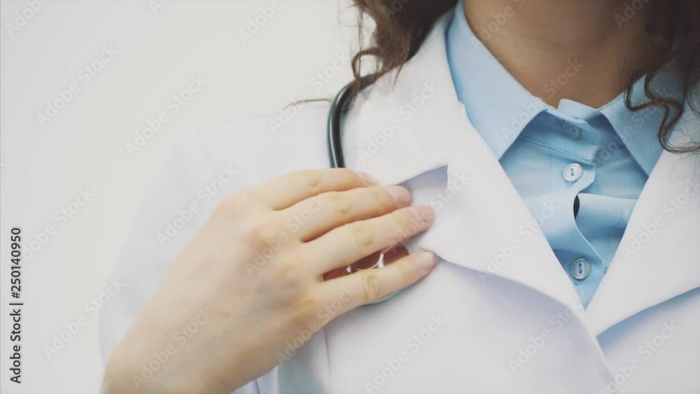 Beautiful young brunette doctor wearing medical clothes on gray background. During this she puts the phonendoscope on her neck. Has beautiful black long hair. Dressed in glasses Slow video. Beautiful young brunette doctor wearing medical clothes on gray background. During this she puts the phonendoscope on her neck. Has beautiful black long hair. Dressed in glasses Slow video.