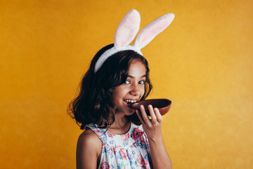 Cute little child wearing bunny ears on Easter day on color background. Girl eating chocolate easter egg