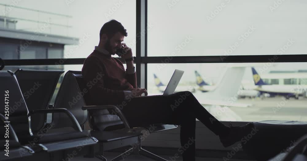 Happy with a good mood businessman working on his laptop and speaking on his phone sitting on the chair in airport , he waiting for the flight , to get boarding