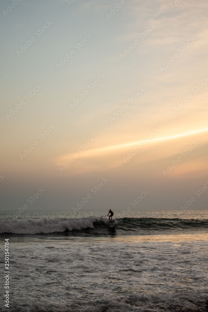 A stand up surfer is riding a wave alone during sunset in Bali, Indonesia A stand up surfer is riding a wave alone during sunset in Bali, Indonesia