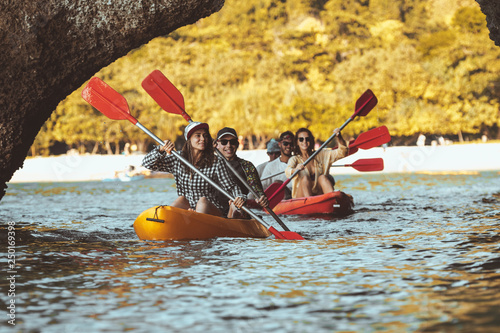 Group of happy friends walks by kayaks under big rocks in the sea. Kayaking or canoeing travel photo with group of peoples