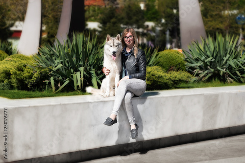 A grey Siberian husky male dog is sitting with young caucasian blond haired girl on marble floor. A woman wears black glasses, grey jeans, black jacket. There're many iris plants around them. © Rabinger