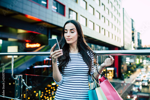Beautiful stylish young woman is using smart phone with shopping bags in hands after mall outdoors