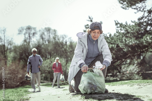 Φωτογραφία Careful old woman in grey jacket putting trash into green packet
