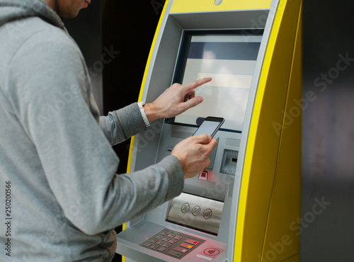 Close up of male hand entering on screen at an ATM. Finger about to press and holding smartphone in other hand. Automated teller machine. 