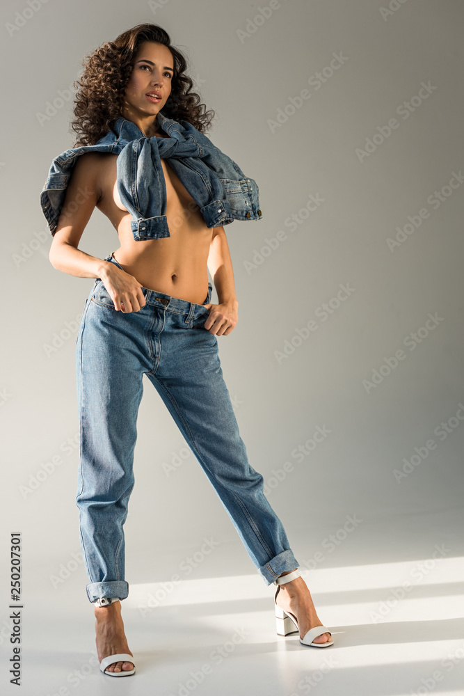 sexy naked woman with curly hair covering breast with denim jacket while  standing in blue jeans on grey background Stock Photo | Adobe Stock