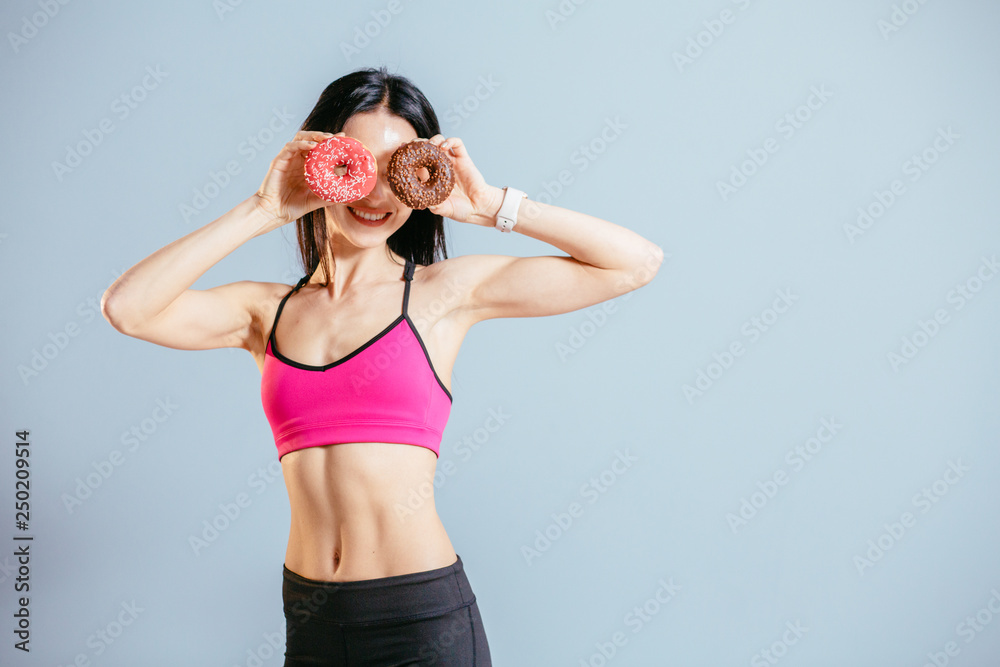 Excited playful sports woman in pink top holding fresh pink and chocolate donuts with powder ready to enjoy sweets. Portrait of attractive brunette slim female in having fun with sweet-stuff.