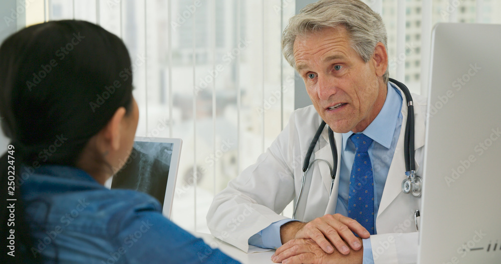 Male Caucasian doctor talking to female patient about neck injury with x-ray on screen. Woman listening to her health care provider explain spinal treatment